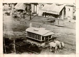 A historical black-and-white photograph of a horse-drawn streetcar traveling on tracks through an urban area. The streetcar has a rounded roof and windows, and two horses are harnessed at the front. In the background, there are storefronts with awnings, pedestrians, and additional horse-drawn carriages.