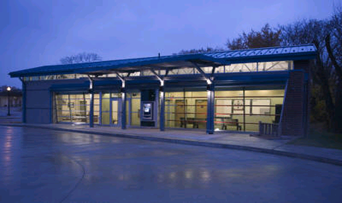 A night picture of the Phyllis Beyers Alliance Transit Center. A SARTA bus station with large glass windows and a metal roof, lit up in the evening. A paved area is in front, with trees in the background.