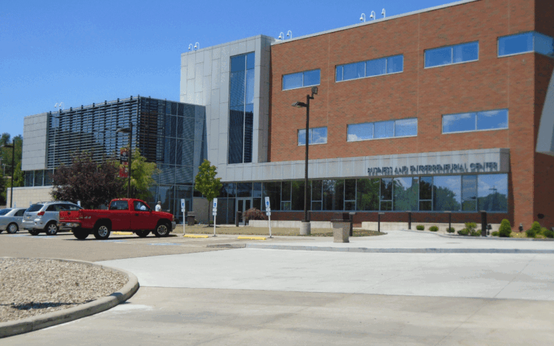 The picture of this Modern multi-story building with a combination of brick and glass architecture. represents the Business Center at Stark State Campus. A parking lot with cars, including a red pickup truck, is visible in the foreground, along with a landscaped area.