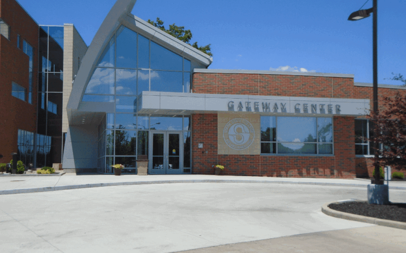 A picture of the Gateway Building at Stark State Campus, the modern glass and brick architecture, features a curved structural design at the entrance. A driveway and streetlamp are visible in the foreground under a clear blue sky.