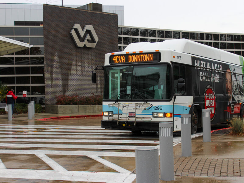 CLEVELAND ROUTE 4- SARTA bus number 1296 parked outside a VA hospital building with a large VA logo on the brick facade. The bus displays the route number and destination '4CVA DOWNTOWN' on its front sign. The bus has an advertisement on its side featuring a personal injury law firm. A pedestrian in a red jacket walks towards the entrance on a rainy day, with wet pavement and a pedestrian crossing visible in the foreground.
