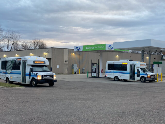 Two white SARTA paratransit buses with blue stripes are parked at a fueling station with a green and white 'Clean Energy' canopy. The SARTA Garage is sitting in the background.