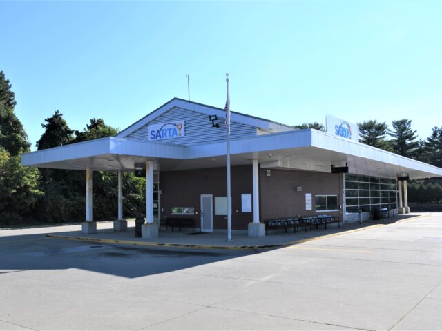 An outside picture of the Belden Village Transit Center on Whipple-SARTA transit center featuring a covered waiting area, seating, and a modern design. The station has a blue SARTA logo on the roof, a glass garage door, and is surrounded by trees under a clear sky
