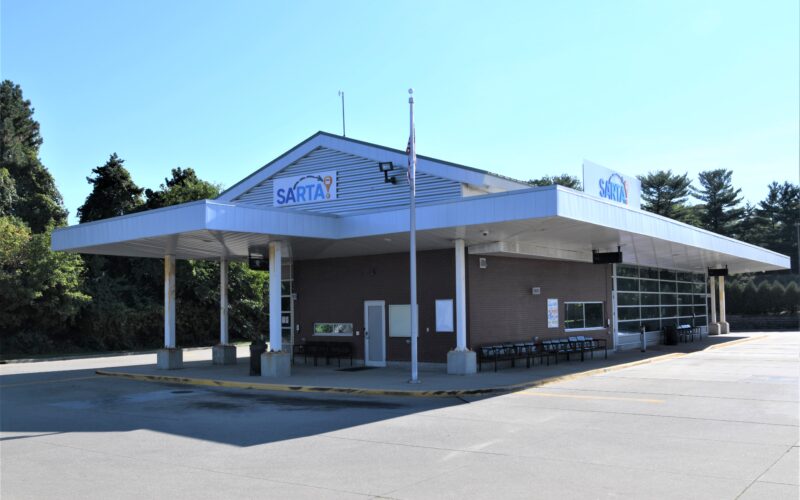 An outside picture of the Belden Village Transit Center on Whipple-SARTA transit center featuring a covered waiting area, seating, and a modern design. The station has a blue SARTA logo on the roof, a glass garage door, and is surrounded by trees under a clear sky
