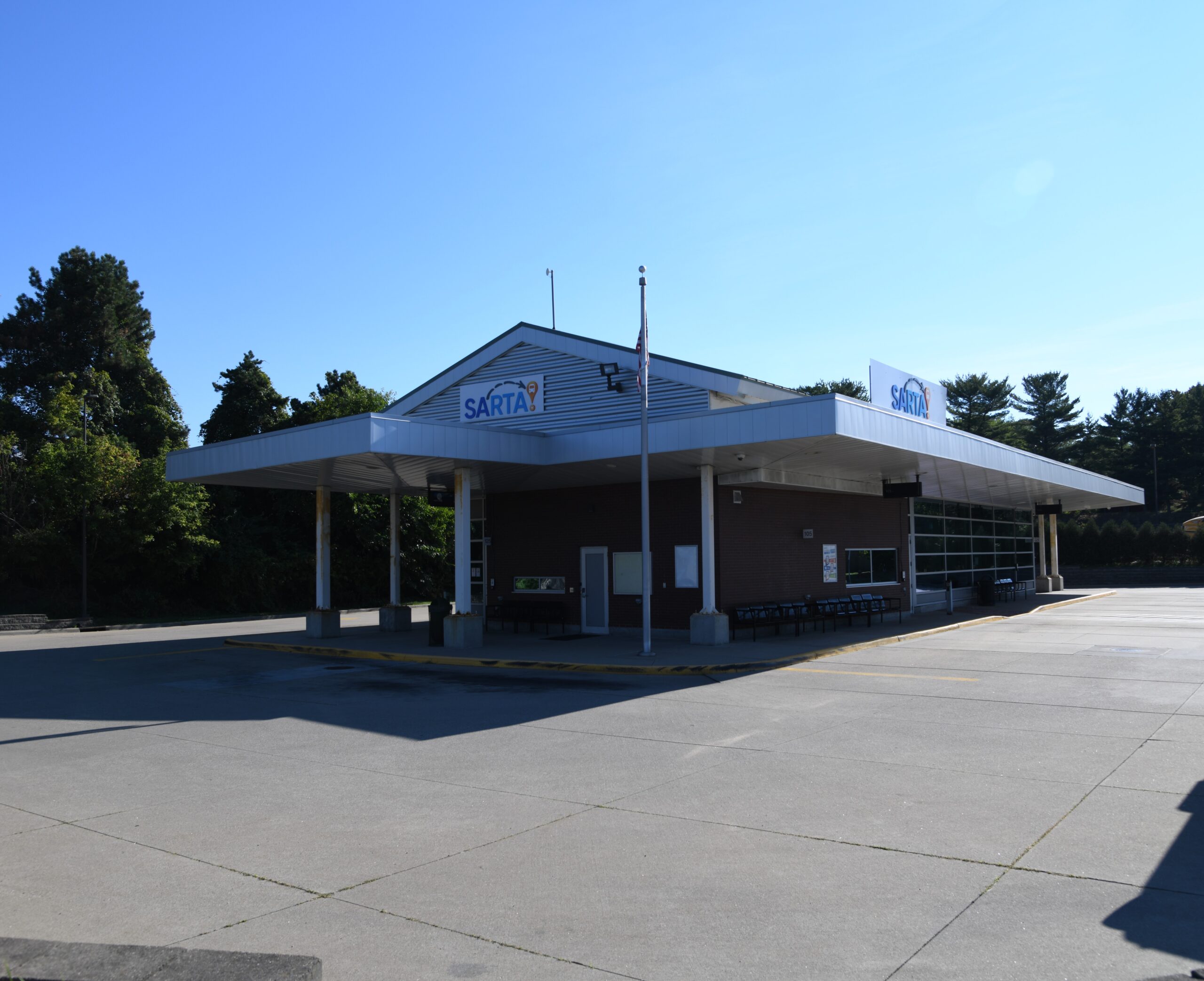 An outside picture of our Belden Village Transit Center. featuring a brick building with a large covered waiting area. The station has a wide concrete lot and is surrounded by trees under a clear blue sky.