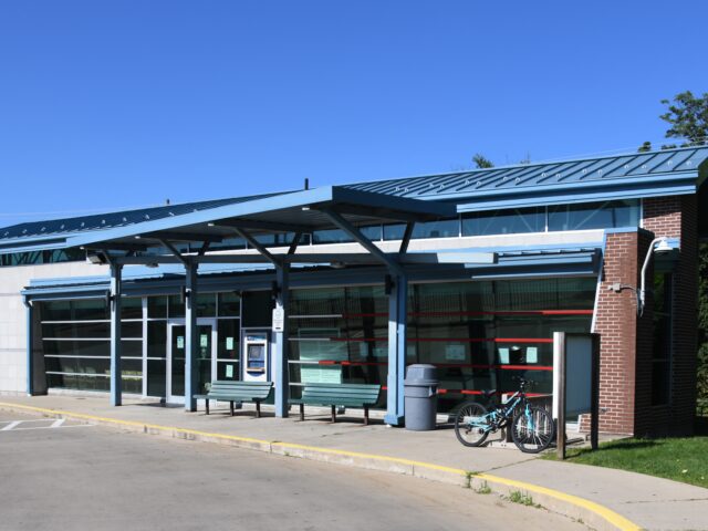 An outside picture of the Phyllis Beyers Alliance Transit Center. This transit center with a blue metal roof and glass-paneled walls, featuring a covered waiting area with benches. A bicycle is parked outside, and the building is surrounded by trees under a clear blue sky.