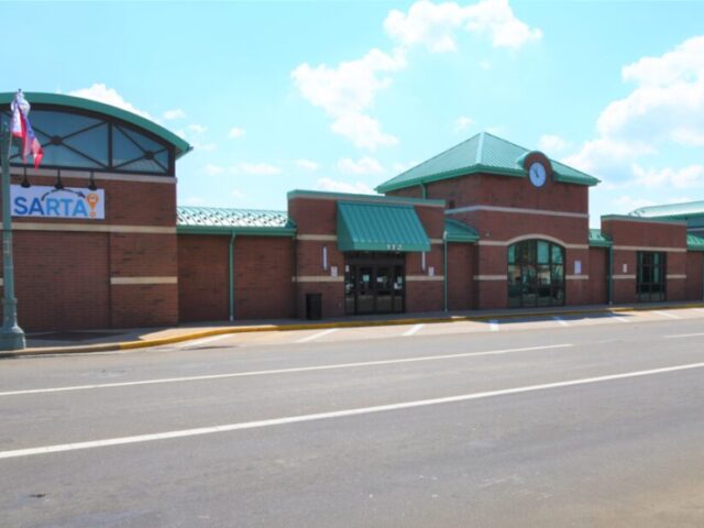 Outside picture on the Downtown Cornerstone Transit Center-Brick transit center with green roof, large arched windows, with SARTA banner. The building has a classic design and sits along a wide street under a bright sky.