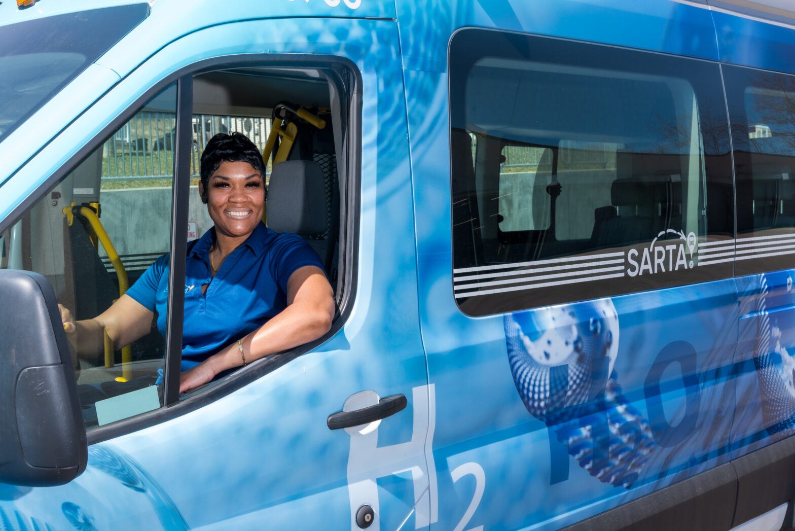 SARTA Bus Driver wearing a blue uniform sits in the driver’s seat of a hydrogen-powered paratransit van smiling. The vehicle has a blue 'H₂O' zero-emissions wrap