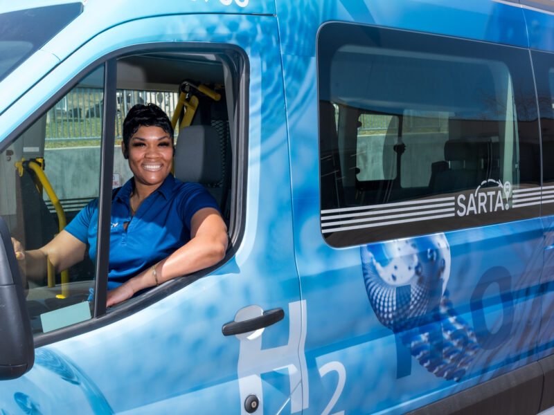 SARTA Bus Driver wearing a blue uniform sits in the driver’s seat of a hydrogen-powered paratransit van smiling. The vehicle has a blue 'H₂O' zero-emissions wrap