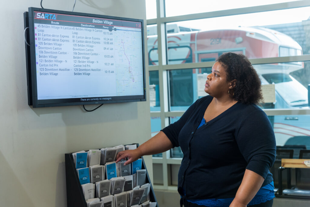 A woman wearing a dark blue cardigan and blue top is standing inside a SARTA bus station, pointing at a brochure rack filled with transit schedules. Above her, a digital screen displays real-time bus arrival times for various routes.