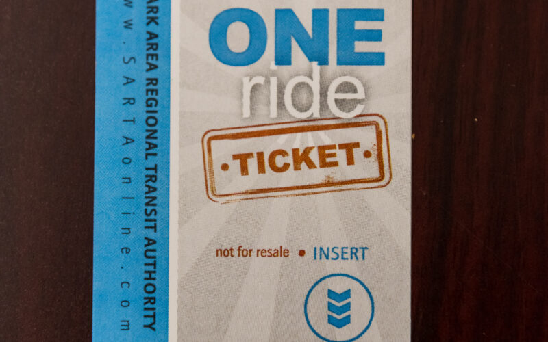 SARTA one-ride ticket featuring a blue and white design with the SARTA logo at the top. The words 'ONE ride TICKET' are prominently displayed in bold lettering. The left side of the ticket has a vertical blue stripe with text, and an arrow at the bottom indicates the correct direction for inserting the ticket. The ticket is placed on a dark wooden surface