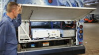 SARTA Technician showing the open rear compartment of a Hydrogen Fuel Cell bus, revealing electrical and mechanical components. The bus has a blue-themed exterior, and various wiring and equipment are visible inside the compartment.