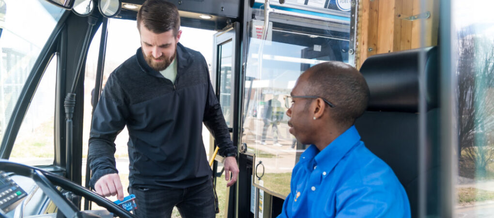 Picture of a rider boarding the bus- Bus driver in a blue shirt sitting at the driver's seat, looking at a male passenger in a black jacket who is boarding the bus and tapping his fare card on the payment reader. The bus interior and a reflection of the outside environment are visible through the windshield.