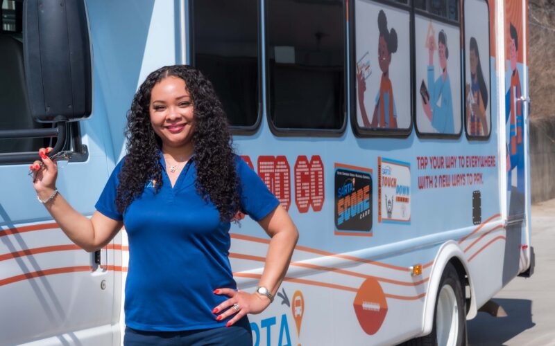 Smiling female SARTA Driver, wearing a blue polo shirt, stands next to a SARTA paratransit bus holding onto the side mirror. The bus features colorful advertisements with illustrated characters promoting public transportation services.