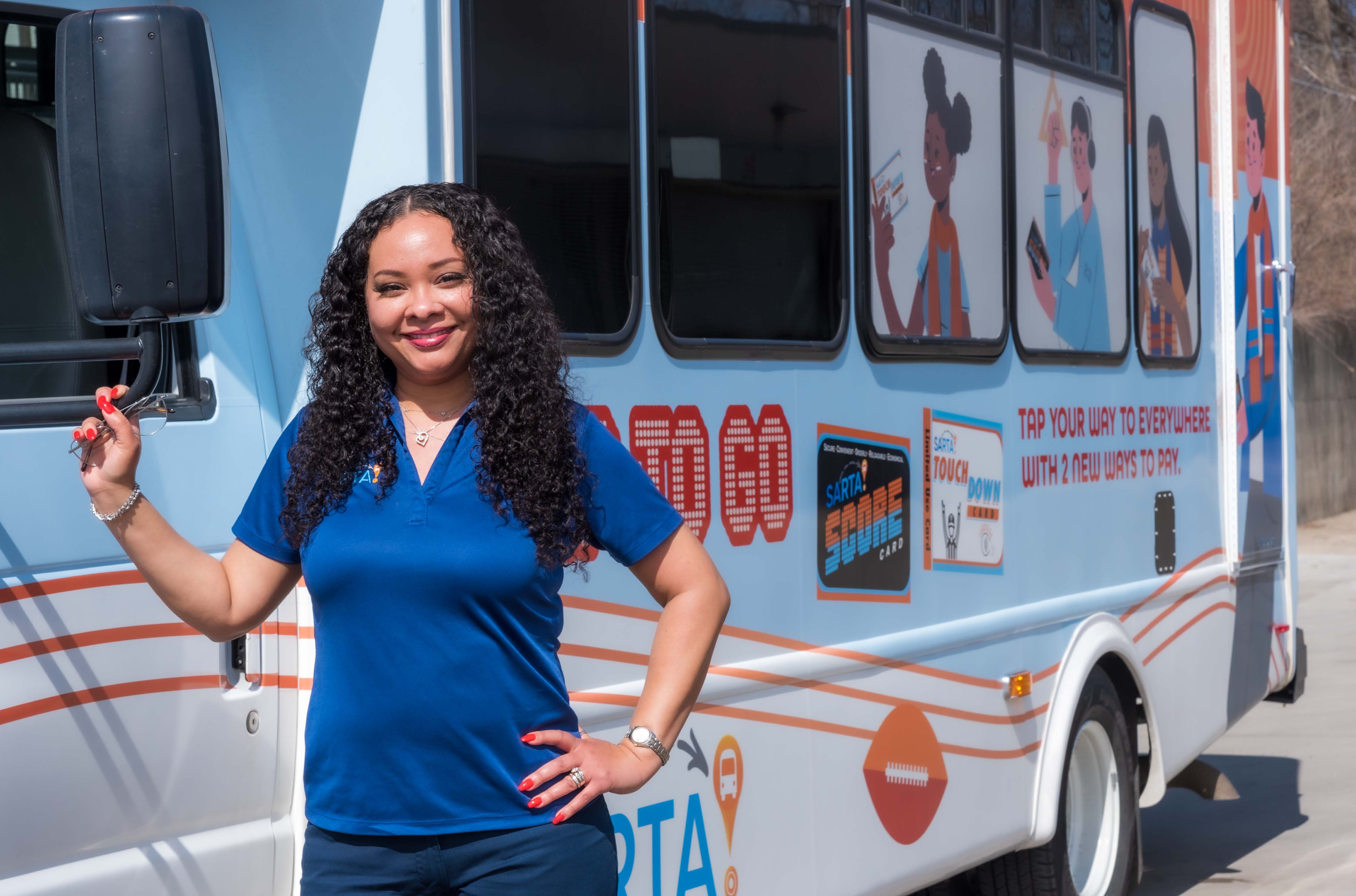 Smiling female SARTA Driver, wearing a blue polo shirt, stands next to a SARTA paratransit bus holding onto the side mirror. The bus features colorful advertisements with illustrated characters promoting public transportation services.