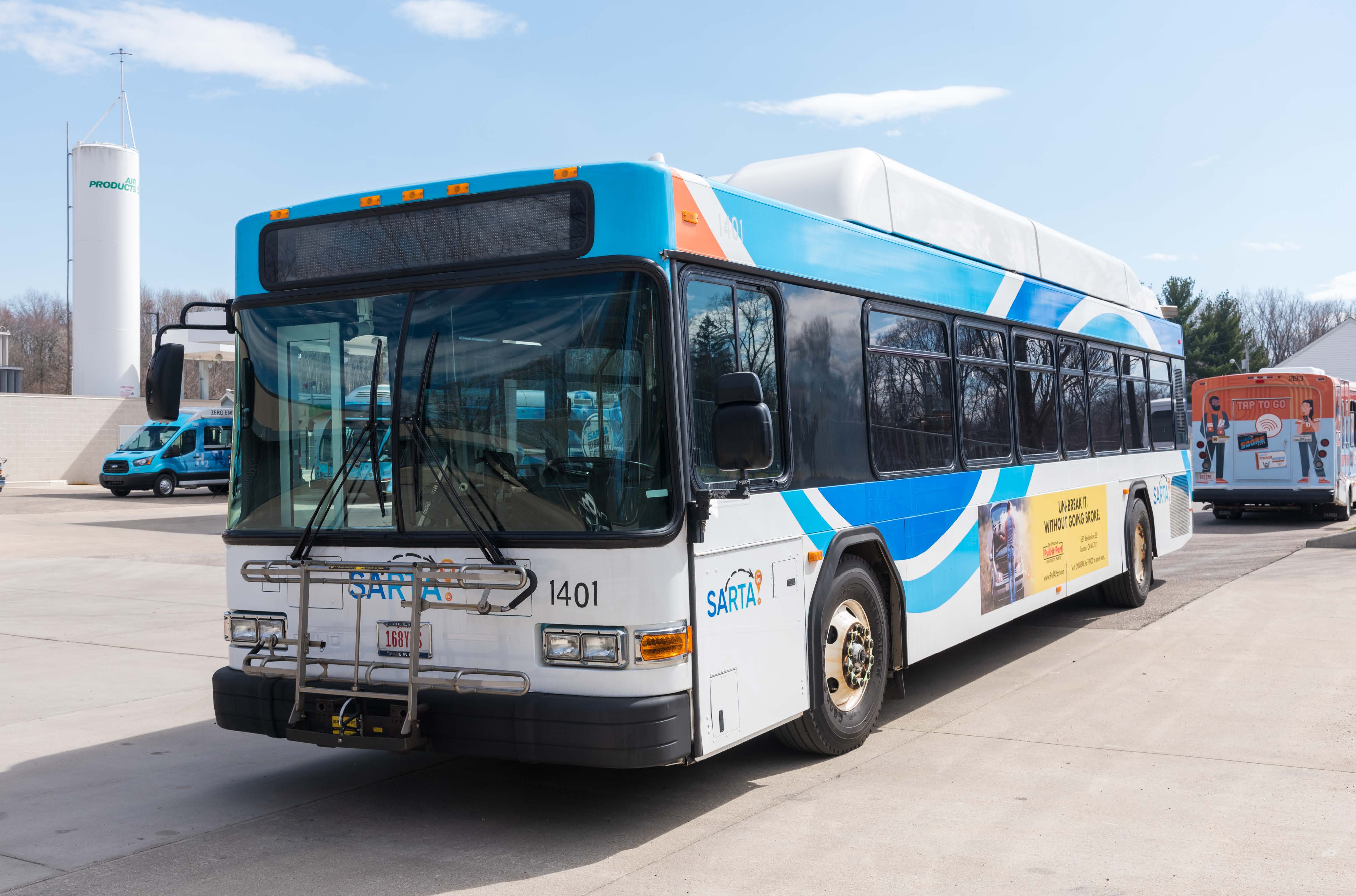 SARTA CNG Bus- A SARTA Compressed Natural Gas (CNG) bus with a blue, white, and orange design is parked in a lot under a partly cloudy sky. The bus has a bike rack on the front. In the background, there are other SARTA vehicles and hydrogen fueling station.