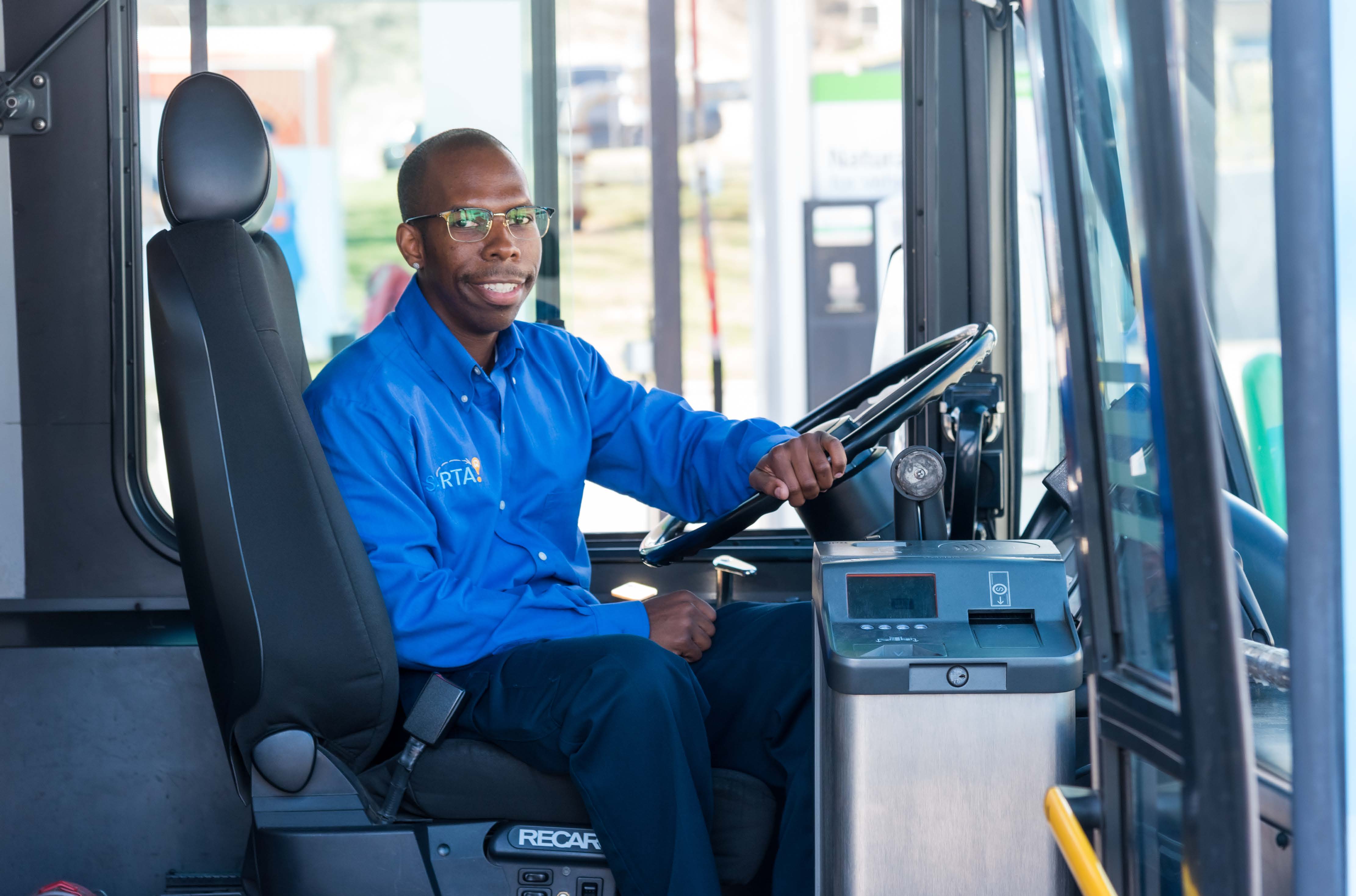 SARTA Driver on a Fixed Route Bus- A male SARTA bus driver wearing a blue uniform sits in the driver’s seat, smiling while holding the steering wheel. The interior of the bus and part of the dashboard are visible, with a fueling station seen in the background through the windshield.