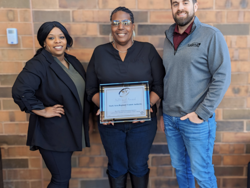 image features members of the SARTA Marketing Team posing with an AdWheel Award. The setting appears to be inside a SARTA facility, with the organization's branding visible in the background. The AdWheel Awards, presented by the American Public Transportation Association (APTA), recognize excellence in marketing and communications in the transit industry.