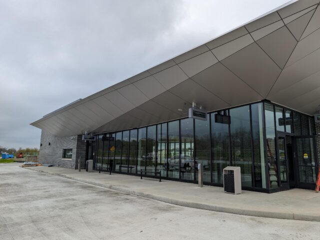 New Massillon Transit Center- A modern transit center with an angular metal roof and large glass windows, creating a sleek and contemporary design. The building is surrounded by a clean concrete area under an overcast sky