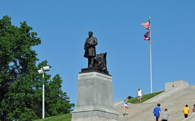 Outdoor scene showing a bronze statue of President William McKinley on a stone pedestal at the top of wide concrete steps, with several people walking on the steps, a U.S. flag and an Ohio state flag flying on poles behind it, green grass and trees to the side, and a clear blue sky overhead.