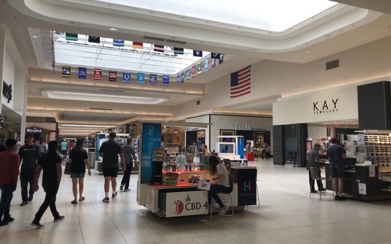 Interior of a shopping mall with a wide central walkway, shoppers walking past kiosk displays, and storefronts including Kay Jewelers and Zales, with banners hanging from the ceiling and an American flag displayed above.