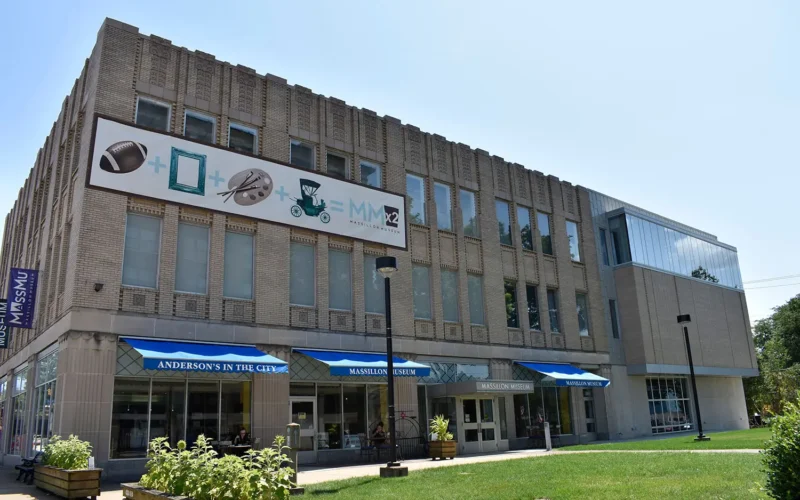 Exterior view of the Massillon Museum, a multi‑story stone building with vertical architectural details, large front windows, and a black decorative street clock in a grassy area in front, with trees and a clear sky nearby.
