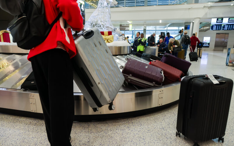 Baggage claim area in an airport showing several suitcases on a moving carousel, with travelers standing nearby holding or pulling rolling luggage inside a bright terminal.